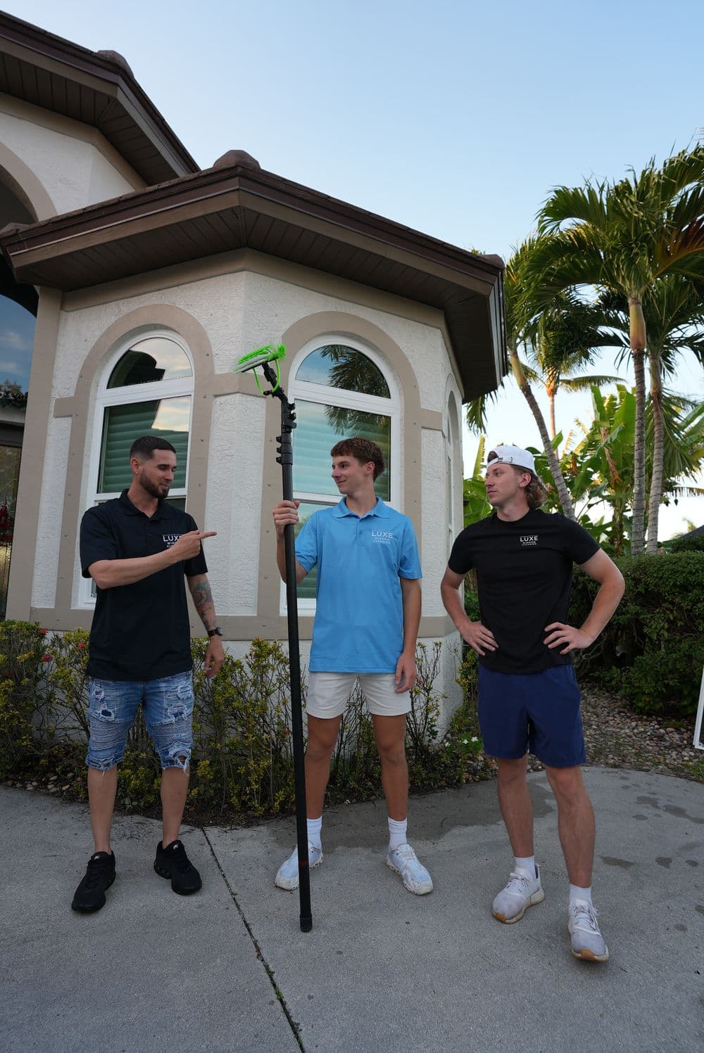 Three people discussing a green cleaning tool outside a modern home with palm trees.