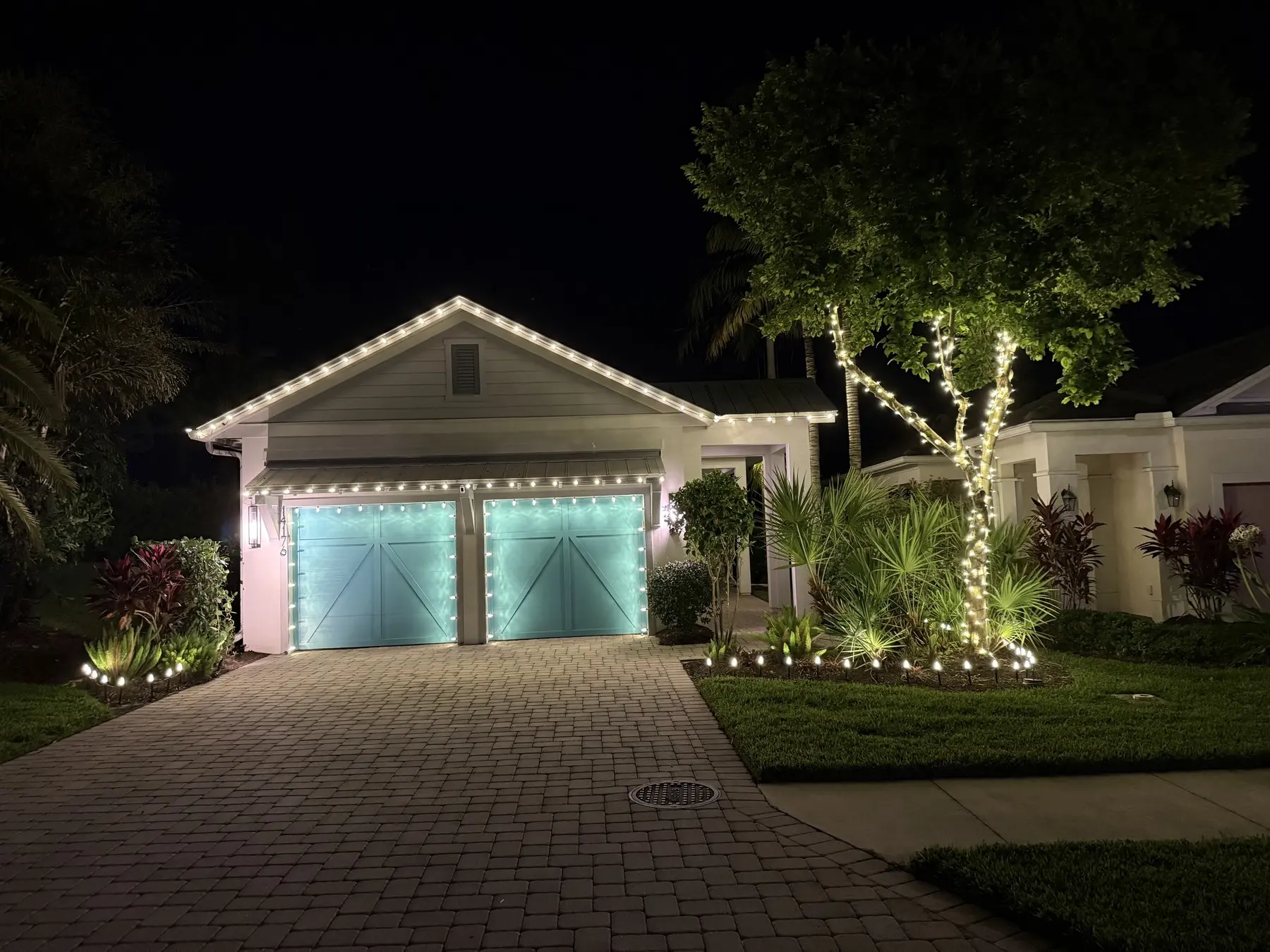 Warm white roofline lighting installed above teal garage doors with a wrapped tree
